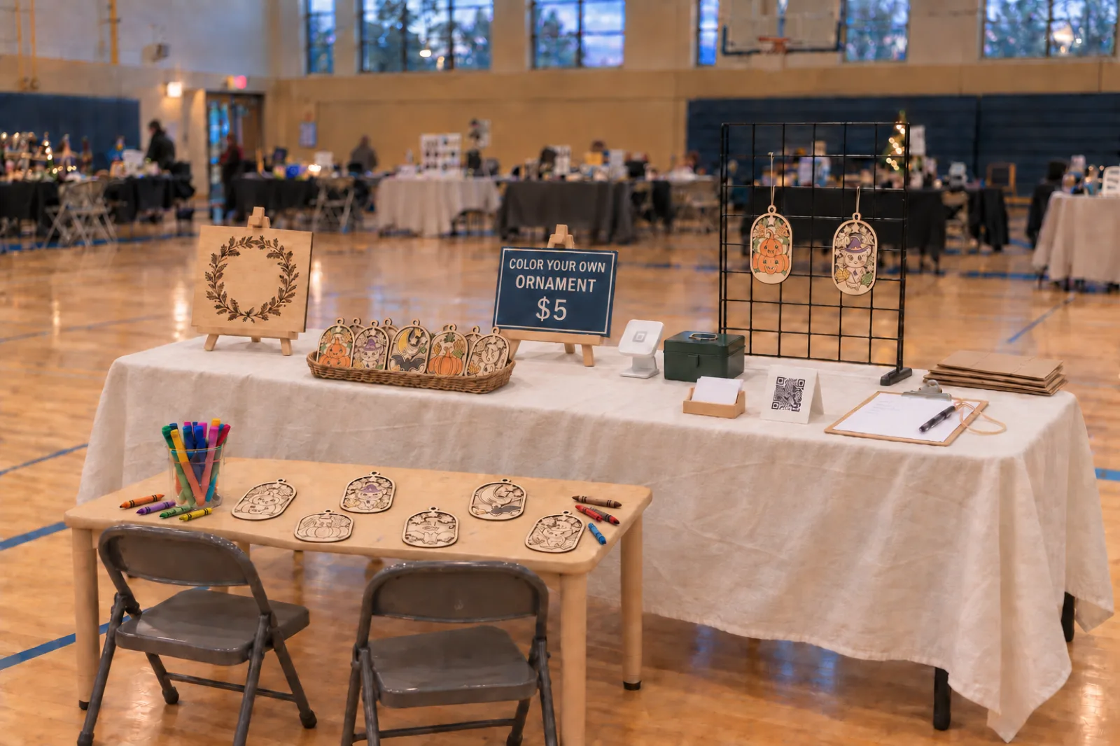 A craft show table set up in a school gym: a colour-your-own ornament station on a low kids' table, a $5 sign, engraved ornaments on a gridwall and an order clipboard